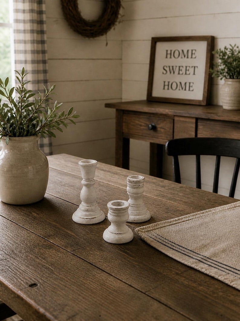 Peut inclure: Une table en bois rustique pr&eacute;sente trois bougeoirs blancs patin&eacute;s. Un vase avec de la verdure et une enseigne encadr&eacute;e avec l'inscription "HOME SWEET HOME" sont &eacute;galement visibles. L'ensemble &eacute;voque une ambiance de ferme chaleureuse.
