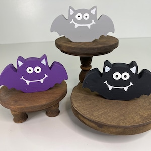 May include: Three wooden bat decorations with painted faces and fangs. The bats are purple, gray, and black. Each bat is displayed on a small wooden pedestal.