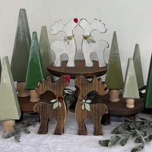 May include: Wooden Christmas decorations featuring reindeer and trees. Two pairs of reindeer are kissing, one pair is white, the other is brown. The trees are painted in shades of green and gray. The reindeer have red noses and decorative accents.