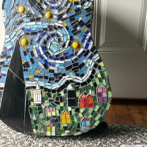 K&ouml;nnte beinhalten: Eine Mosaikgitarre mit einem Sternennacht-Design. Die Kunstwerke verwenden blaue, schwarze, gelbe und gr&uuml;ne Fliesen, um einen wirbelnden Nachthimmel, ein Dorf und einen Berg darzustellen. Der Korpus der Gitarre ist schwarz.