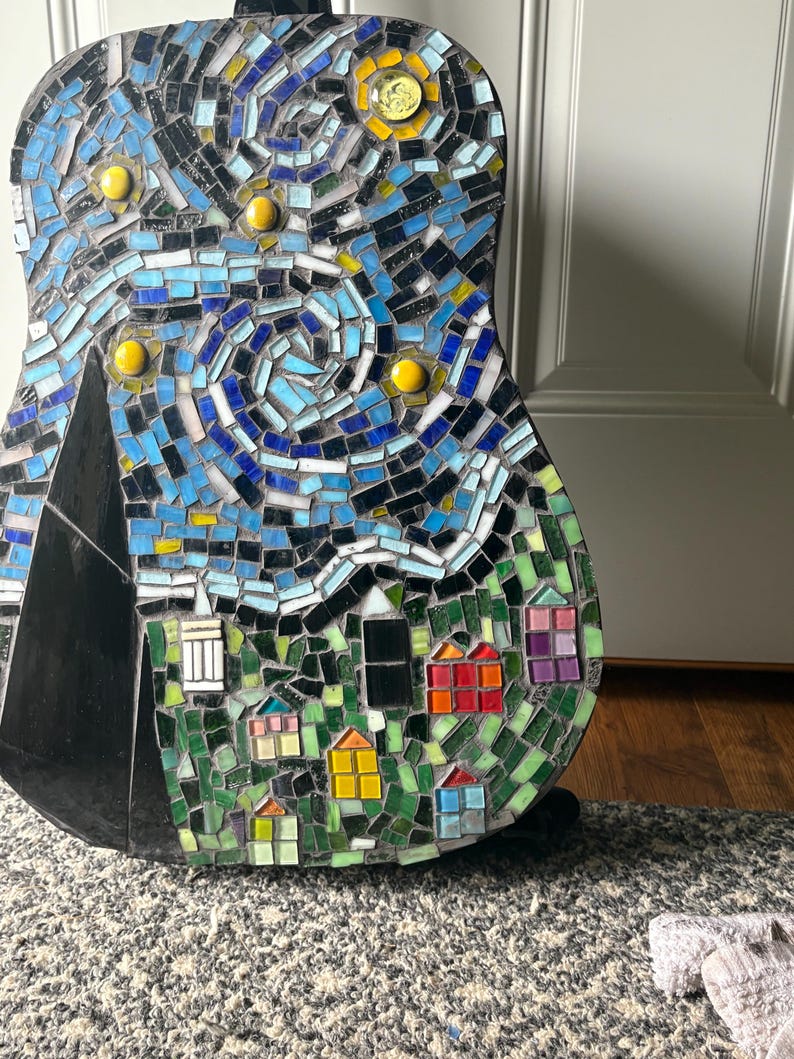 K&ouml;nnte beinhalten: Eine Mosaikgitarre mit einem Sternennacht-Design. Die Kunstwerke verwenden blaue, schwarze, gelbe und gr&uuml;ne Fliesen, um einen wirbelnden Nachthimmel, ein Dorf und einen Berg darzustellen. Der Korpus der Gitarre ist schwarz.