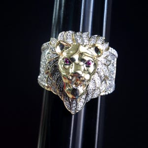 May include: A gold and silver lion ring with ruby eyes. The lion's head is gold, and the mane and band are encrusted with clear gemstones. The ring is displayed against a black background.