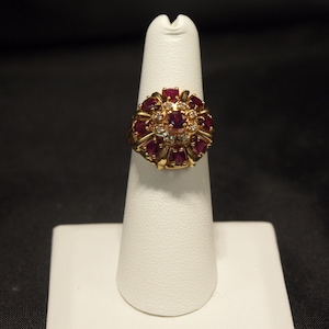 May include: A gold ring featuring a central cluster of small clear stones surrounded by a halo of larger, deep red, oval-cut stones. The design is arranged in a circular, floral-like pattern with a polished gold band and setting.