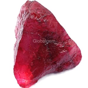 Op de afbeelding: Een ruwe, ongeslepen robijn edelsteen met een dieprode kleur. Het oppervlak van de steen heeft een textuur, met zichtbare facetten en onregelmatigheden. De tekst "Globalgem" is zichtbaar op de steen.