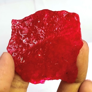 Op de afbeelding: Een levendige rode edelsteen met een getextureerd oppervlak. De onregelmatig gevormde steen wordt omhoog gehouden, waarbij de diepe kleur en natuurlijke kenmerken worden getoond. De edelsteen lijkt een ruw, ongeslepen exemplaar te zijn, mogelijk een robijn of een vergelijkbaar mineraal.
