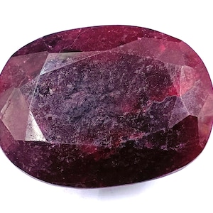 Può includere: Una gemma di rubino sfaccettata di forma ovale. La gemma ha un colore rosso intenso con variazioni di tonalità e inclusioni interne visibili. Il taglio della pietra rivela molteplici sfaccettature, esaltandone la brillantezza. Il rubino è su sfondo bianco.