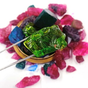 May include: A close-up of a green gemstone being held by tweezers. The gemstone is surrounded by other gemstones in various colors, including red, blue, and purple.