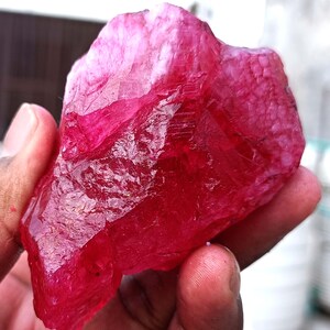 May include: A raw, uncut ruby gemstone in a deep red colour. The surface has a textured appearance with areas of lighter pink and white. The gemstone is held in a hand, showcasing its natural, raw form. Suitable for jewellery making or gemstone collectors.