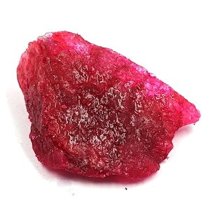 May include: A raw, unpolished ruby gemstone with a deep, vibrant red color. The stone features a jagged, irregular shape with a textured, crystalline surface that catches light, displaying varying shades of intense crimson and magenta.