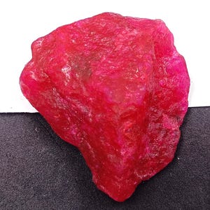Könnte beinhalten: Ein rauer, ungeschliffener Rubin-Edelstein. Der Rubin hat eine tiefe, leuchtende rote Farbe mit einer strukturierten Oberfläche. Der Edelstein wird vor einem weißen und schwarzen Hintergrund präsentiert.