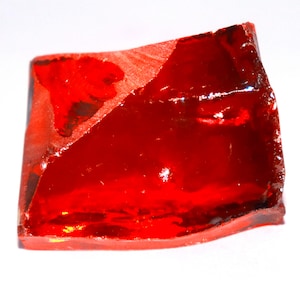 Op de afbeelding: Een doorschijnend, dieprood glasfragment met een onregelmatige, hoekige vorm. Het oppervlak vertoont een mix van gladde, gepolijste facetten en ruwe, getextureerde randen die het licht vangen en levendige tinten van robijnrood en karmozijn creëren.