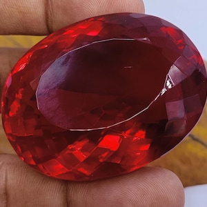 May include: A large, faceted, oval-shaped gemstone with a deep red hue. The gem is held in a hand, showcasing its polished surfaces and internal reflections. The cut of the stone creates multiple facets, enhancing its brilliance.