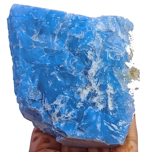 May include: A large, raw chunk of vibrant blue mineral with a glassy, translucent texture. The surface features irregular, jagged edges and deep ridges, with patches of white, crystalline inclusions visible along the right side and crevices.
