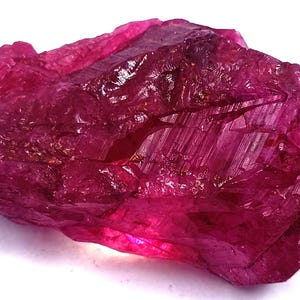 May include: A raw, unpolished gemstone with a deep magenta and fuchsia color. The stone features a jagged, crystalline structure with sharp, angular facets and a translucent quality that allows light to pass through its irregular, textured surface.