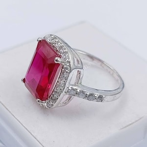 Puede incluir: Un anillo de plata con una gran piedra roja de talla esmeralda rodeada de piedras pequeñas y transparentes.