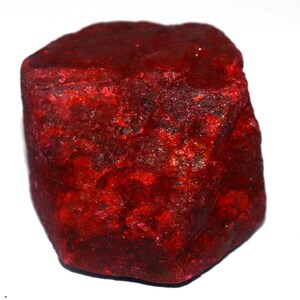Puede incluir: Una piedra preciosa de rubí en bruto y sin tallar con un color rojo intenso y saturado. La piedra presenta una superficie rugosa e irregular con texturas cristalinas naturales, bordes afilados y tonos variables de carmesí y escarlata.