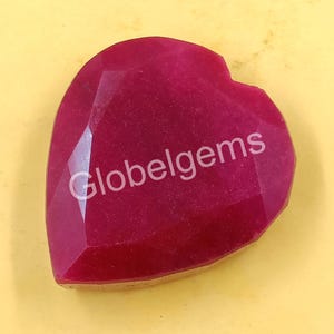 Può includere: Una gemma sfaccettata a forma di cuore, di colore magenta intenso. La superficie riflette la luce, mostrando molteplici sfaccettature. Il testo "Globelgems" è stampato sulla gemma. Lo sfondo è giallo chiaro.