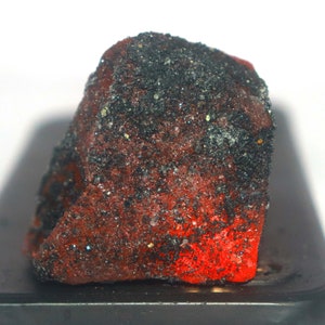Op de afbeelding: Een ruw mineraal exemplaar met een dieprode, doorschijnende basiskleur bedekt met een dichte, getextureerde laag van donkerzwarte, korrelige deeltjes. Het oppervlak vertoont een ruwe, onregelmatige kristalstructuur met kleine reflecterende punten.