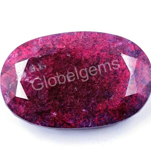 May include: A deep red oval gemstone with a faceted cut and a purple hue. The gemstone has a rough, textured surface.