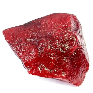 Puede incluir: Un espécimen mineral en bruto de forma irregular con un color rojo intenso y vibrante. La superficie de la piedra presenta una textura cristalina rugosa con motas metálicas de color dorado incrustadas a lo largo de los bordes dentados.