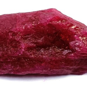 May include: A raw ruby gemstone, displaying a rich, deep red colour. The stone's surface is textured, revealing crystalline formations and variations in colour. The ruby is presented against a white backdrop.