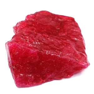 Könnte beinhalten: Ein roher, ungeschliffener Rubin-Edelstein mit tiefroter Farbe. Die Oberfläche hat ein strukturiertes Aussehen mit sichtbaren Graten und Farbtonvariationen. Der Edelstein ist vor einem weißen Hintergrund platziert, um seine natürliche Form und Farbe hervorzuheben.