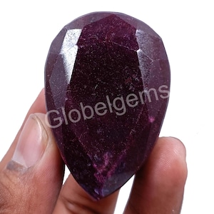 May include: A deep purple gemstone with a faceted pear shape. The gemstone is held in a hand, and the text "Globelgems" is visible in the image.