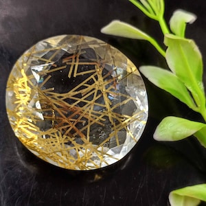 May include: A clear quartz crystal with gold inclusions. The crystal is faceted and has a round shape. It is set against a black background.