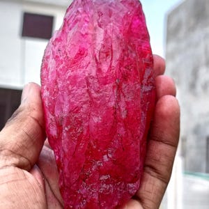 May include: A large, raw ruby gemstone with a deep red color. The rough-cut stone has a textured surface and is held in a hand, showcasing its natural form and vibrant hue. The ruby is a gemstone.