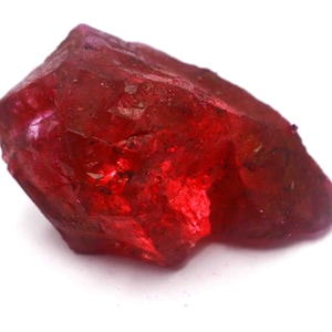 May include: A raw, unpolished gemstone with a deep, vibrant red color. The stone features an irregular, jagged shape with multiple natural facets and crystalline textures that catch the light, creating varying shades of crimson and scarlet throughout.