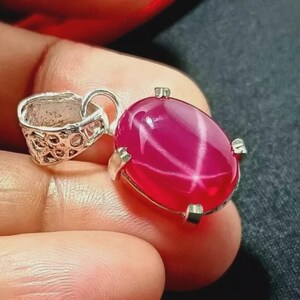 May include: A silver pendant featuring a large, oval ruby gemstone. The ruby displays a star effect and is set in a silver bezel. The pendant's bail is silver and has a decorative design.