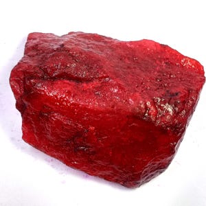 Może przedstawiać: Surowy, nieoszlifowany kawałek jaskrawoczerwonego minerału o krystalicznej, półprzezroczystej teksturze. Powierzchnia posiada nieregularne fasetki, głębokie szkarłatne odcienie oraz naturalne wypukłości, które odbijają światło.