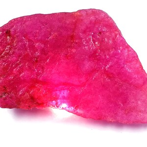 May include: A rough-cut, vibrant pink ruby gemstone. The natural stone has an irregular shape and a textured surface, showcasing its raw, unpolished state. The ruby's color ranges from deep magenta to lighter pink hues.