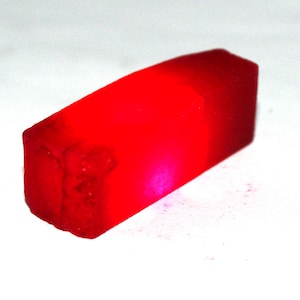May include: A rectangular, translucent red gemstone with a rough, unpolished texture. The stone exhibits a vibrant, glowing crimson hue throughout its crystalline structure, with a slightly darker, saturated red tone concentrated in the center.