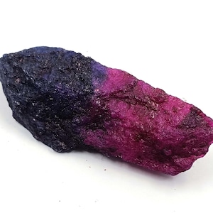 May include: A raw, elongated mineral specimen featuring a distinct color gradient. One end is a deep, dark navy blue with a rough, textured surface, transitioning into a vibrant, saturated magenta pink on the opposite end.