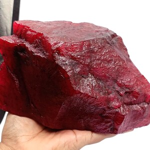 May include: A large, uncut ruby gemstone with a deep red hue. The raw mineral has a textured surface and an irregular shape, held in a hand against a white background. The ruby appears to be natural and unpolished.