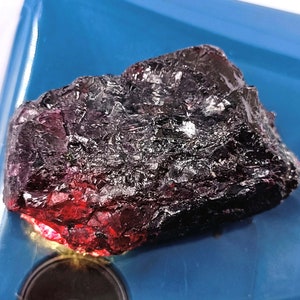 May include: A large, dark purple and red crystal with a rough, textured surface. The crystal is sitting on a blue surface.