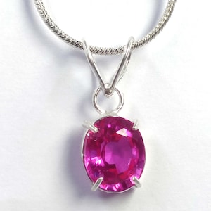 Könnte beinhalten: Eine silberne Anhängerkette mit einem leuchtenden, oval geschliffenen rosa Edelstein. Der Edelstein ist in einer silbernen Fassung gefasst und hängt an einer zarten, strukturierten Silberkette. Die leuchtend rosa Farbe des Steins ist der Blickfang des Schmucks.