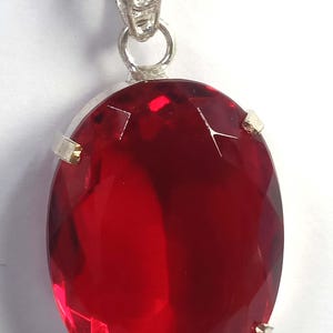 May include: A silver pendant necklace featuring a large, oval-cut, deep red gemstone. The gemstone is set in a silver bezel with small gold-colored accents. The chain is a delicate silver link.