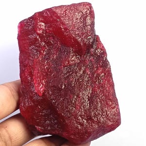 Puede incluir: Una gema de rubí grande, cruda y sin cortar con un color rojo profundo y saturado. La piedra presenta una textura cristalina rugosa con facetas naturales y bordes irregulares, mostrando varios tonos de carmesí en toda su superficie.
