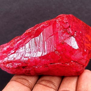 May include: A raw, uncut ruby gemstone with a deep red hue. The surface of the stone has a rough, textured appearance with reflective facets. The ruby is held in a hand against a dark background. Gemstone.
