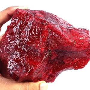 May include: A large, raw, unpolished gemstone with a deep, vibrant red colour. The stone features a rough, crystalline texture with natural facets and irregular edges, displaying a translucent quality that catches light across its uneven surface.