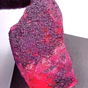 May include: A large, rough, red gemstone with a dark purple hue. The gemstone has a crystalline texture and is irregularly shaped.