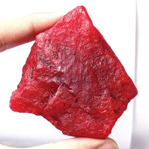 May include: A raw, unpolished gemstone with a vibrant, deep red color. The stone features an irregular, angular shape with a textured, crystalline surface that displays natural facets and dark inclusions throughout its translucent structure.