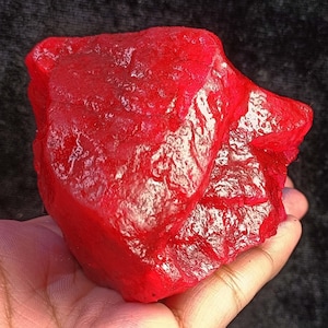 May include: A large, rough-cut, deep red gemstone with a textured surface. The gemstone is held in a hand, showcasing its size and color. The stone has a natural, unpolished appearance, highlighting its raw beauty.