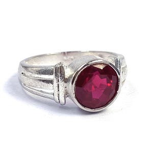 Puede incluir: Un anillo de plata con una piedra preciosa redonda, facetada y de color rojo intenso engastada en un bisel. La banda presenta un diseño texturizado con múltiples ranuras a ambos lados de la piedra central, con un acabado metálico pulido.