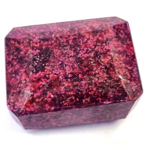 Puede incluir: Una piedra preciosa rectangular con una superficie facetada, que muestra un patrón moteado de color magenta intenso y púrpura oscuro en toda su estructura. La piedra tiene un acabado pulido con bordes afilados y una apariencia granular densa.