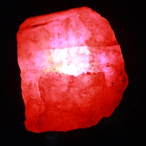May include: A raw, translucent mineral specimen with an irregular, jagged shape. The stone exhibits a vibrant, glowing red hue throughout its crystalline structure, with a brighter, concentrated white light source visible in the center.