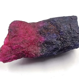 May include: A raw, unpolished mineral specimen featuring a distinct color gradient. The left side is a vibrant, saturated magenta pink, transitioning into a deep, dark navy blue and black on the right side with a rough, textured surface.