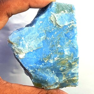 May include: A raw, unpolished chunk of blue opal mineral with a jagged, crystalline texture. The stone displays varying shades of sky blue and translucent white, featuring natural fractures and uneven surfaces throughout its irregular shape.